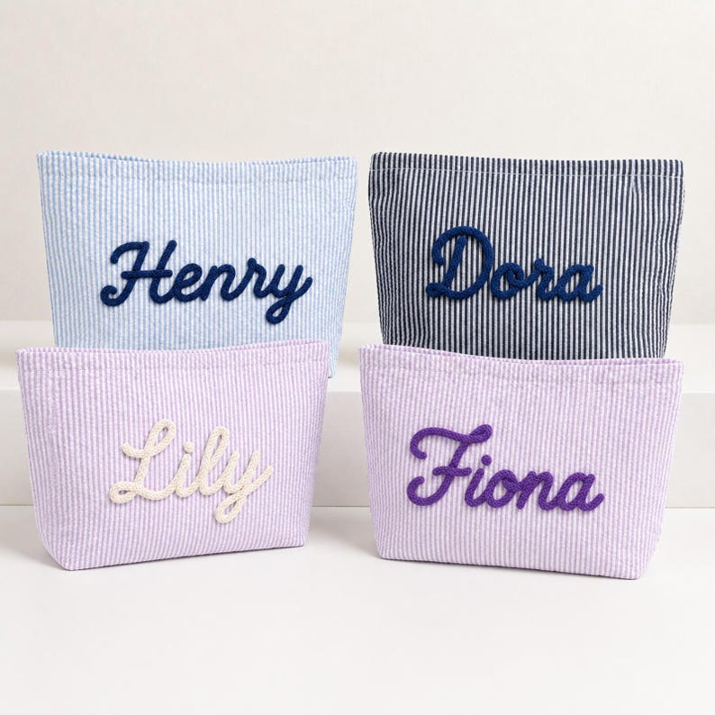 Puede incluir: Cuatro estuches rectangulares de tela con rayas verticales. El estuche superior izquierdo es azul claro con el nombre Henry en azul oscuro. El superior derecho es azul marino con el nombre Dora en marino. El inferior izquierdo es lavanda con el nombre Lily en blanco y el inferior derecho es lavanda con el nombre Fiona en morado.