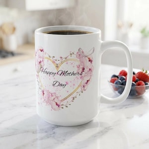 May include: White ceramic mug with a large handle, filled with dark liquid and steam rising. The mug features a heart-shaped design with pink floral accents and the text "Happy Mothers Day". A small bowl of berries sits nearby.