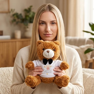Puede incluir: Un oso de peluche marrón con una camiseta blanca y una pajarita negra. La camiseta tiene un gráfico de un ángel y el texto "Hugs Sent From Heaven". El oso está siendo sostenido.