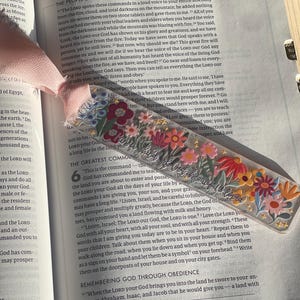 Peut inclure: Un marque-page rectangulaire transparent orné d'un motif floral coloré avec des fleurs rouges, roses, jaunes et bleues, des feuilles vertes et de petites étoiles dorées. Le texte "child of God, beautiful & loved" est écrit en cursive.
