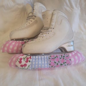 May include: A pair of white ice skates with grey laces and silver heels, each featuring a protective blade cover made of fabric with a patchwork pattern of pink cherries, light blue gingham checks, and pink floral prints on a white background.