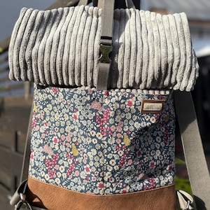 Könnte beinhalten: Ein grauer Cord-Rucksack mit einer floral gemusterten Frontplatte und einer braunen Kunstlederbasis. Der Rucksack hat verstellbare Träger und einen Metall-Schnallenverschluss. Das Etikett lautet "CH DESIGN".