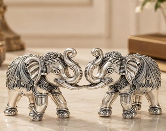 May include: Two silver-toned elephant figurines facing each other, featuring intricate embossed patterns on their bodies, ears, and trunks. The elephants are depicted in a standing position with their trunks curled upward, showcasing detailed metallic textures.