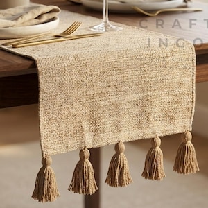 Peut inclure: Un chemin de table rectangulaire en tissage beige avec cinq pompons décoratifs sur le bord inférieur. Il est posé sur une table en bois foncé avec une assiette blanche, un verre à vin transparent et un petit vase avec des feuilles vertes.