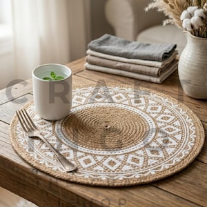 May include: A round, woven jute placemat featuring a white printed geometric pattern. The placemat is placed on a wooden table alongside a white ceramic mug containing a green sprig, a silver fork, and a stack of folded fabric napkins.