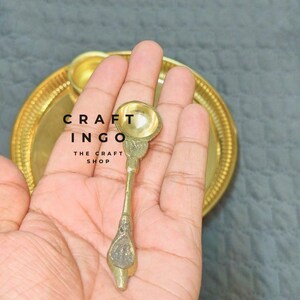 May include: A small, metallic gold-toned spoon with a rounded bowl and a textured handle, positioned in front of a matching circular gold-toned tray. The text CRAFT INGO THE CRAFT SHOP is visible in black lettering on the surface of the tray.