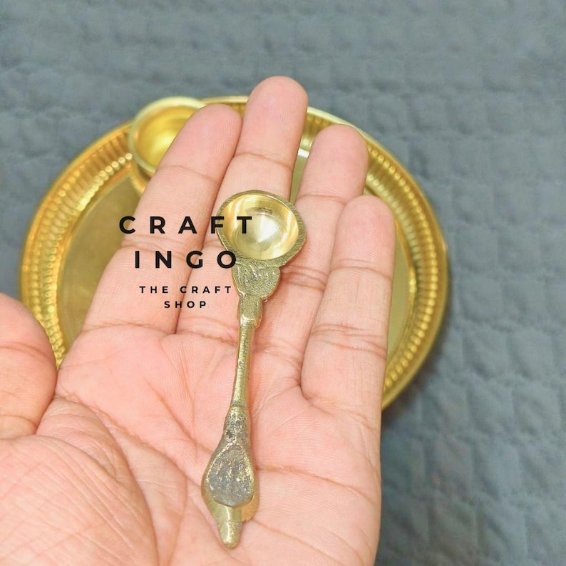 May include: A small, metallic gold-toned spoon with a rounded bowl and a textured handle, positioned in front of a matching circular gold-toned tray. The text CRAFT INGO THE CRAFT SHOP is visible in black lettering on the surface of the tray.