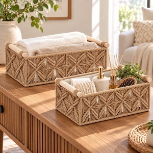 Puede incluir: Dos cestas de almacenamiento rectangulares con un exterior de cuerda de macramé color canela con patrón de diamantes y asas de madera clara. Las cestas contienen toallas blancas dobladas, un dispensador de jabón, un difusor y un cepillo.