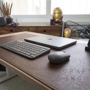 Könnte beinhalten: Eine dunkelbraune Schreibtischunterlage aus Leder mit genähten Kanten, darauf ein graues kabelloses Keyboard, eine graue kabellose Maus und ein geschlossener grauer Laptop. Der Text CRAFT INGO THE CRAFT SHOP steht unten rechts auf der Unterlage.