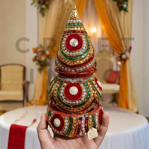 May include: A decorative, multi-layered item with a conical top. It features red, green, and gold embellishments, including sequins and beads, with a central white accent. The base is round. The background shows draped fabric and floral decorations.