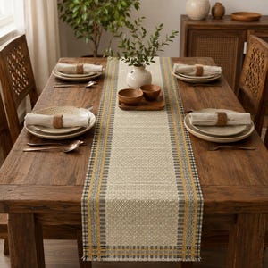 Pode incluir: Um caminho de mesa retangular tecido com um centro bege de padrão geométrico, bordado por riscas verticais em amarelo mostarda, preto e branco. O caminho de mesa está colocado sobre uma mesa de jantar de madeira escura com loiça e um vaso.
