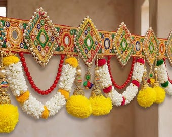 May include: A decorative door hanging featuring a horizontal band with embroidered diamond shapes, mirrors, and red beads. White floral garlands drape below, accented with bright yellow pom-poms and gold-toned bell-shaped ornaments.