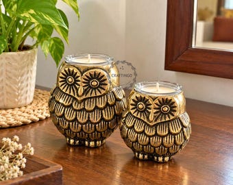 May include: Two gold-colored owl-shaped candle holders with textured feather details and clear glass inserts containing white wax candles. The larger owl is on the left and the smaller owl is on the right. The text CRAFTINGO is visible.