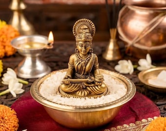 May include: A brass-colored figurine of a seated deity holding a small spoon and a bowl, positioned in the center of a shallow brass bowl filled with white rice grains. The figure features intricate detailing on the crown and traditional attire.