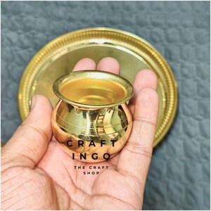May include: A small, polished brass pot with a rounded body and a wide rim, accompanied by a matching circular brass tray. The text CRAFT INGO THE CRAFT SHOP is printed in black on the surface of the pot. The metal has a reflective finish.