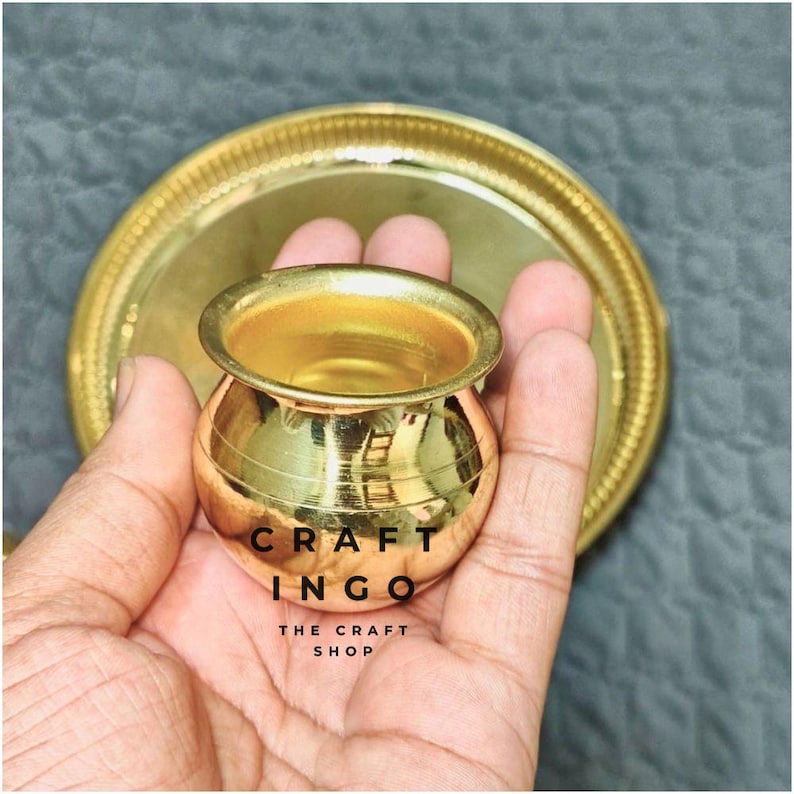 May include: A small, polished brass pot with a rounded body and a wide rim, accompanied by a matching circular brass tray. The text CRAFT INGO THE CRAFT SHOP is printed in black on the surface of the pot. The metal has a reflective finish.