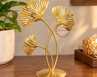 May include: A gold-colored metal sculpture featuring three ginkgo leaf shapes attached to thin stems rising from a circular base. The leaves have a ribbed texture and a metallic finish. A circular logo with the text CRAFTINGO is visible.