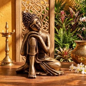 Könnte beinhalten: Eine bronzefarbene Buddha-Statue in sitzender, ruhender Pose, bei der ein Arm auf einem angehobenen Knie ruht. Die Figur zeigt eine texturierte, gelockte Frisur und glatte, fließende Gewänder. Sie ist mittig platziert und hat eine metallische Oberfläche.