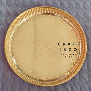 May include: A round, polished gold-tone metal tray featuring an intricate, embossed floral pattern across its surface and a ribbed, textured rim. The text CRAFT INGO THE CRAFT SHOP is printed in black on the right side of the tray's center.