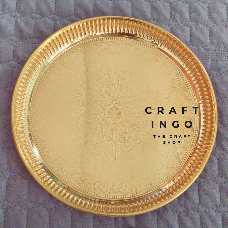May include: A round, polished gold-tone metal tray featuring an intricate, embossed floral pattern across its surface and a ribbed, textured rim. The text CRAFT INGO THE CRAFT SHOP is printed in black on the right side of the tray's center.