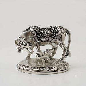 May include: A silver-toned metal figurine depicting a cow and a calf standing on a circular base. The cow features an ornate blanket with an Om symbol on its back. The base has a textured pattern, and the entire piece has a polished finish.