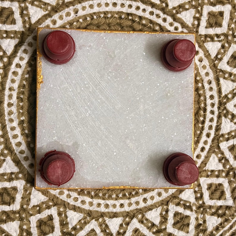May include: A square white stone coaster with a gold-painted edge. The underside features four small, round, dark red rubber feet positioned at each corner to provide elevation and stability on flat surfaces.