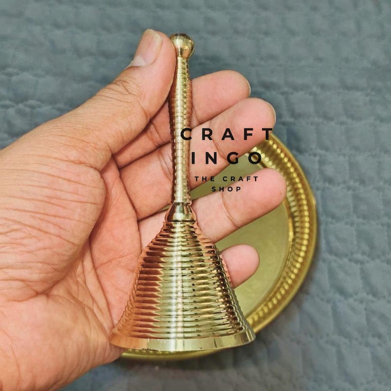 May include: A polished brass hand bell with a ribbed, conical body and a slender, textured handle. The bell is positioned in front of a matching circular brass tray. The text CRAFT INGO THE CRAFT SHOP is visible in the center of the image.