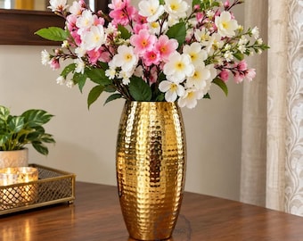 May include: A tall, cylindrical, hammered gold metal vase holds a large arrangement of artificial flowers featuring white and pink blossoms with green leaves. The vase has a reflective, textured surface and sits upright.