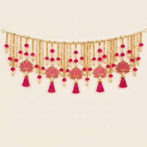 May include: A decorative wall hanging featuring a horizontal string of cream-coloured beads interspersed with vibrant pink pom-poms. Five pink lotus-shaped cutouts are suspended from the main strand, each finished with a matching pink tassel.
