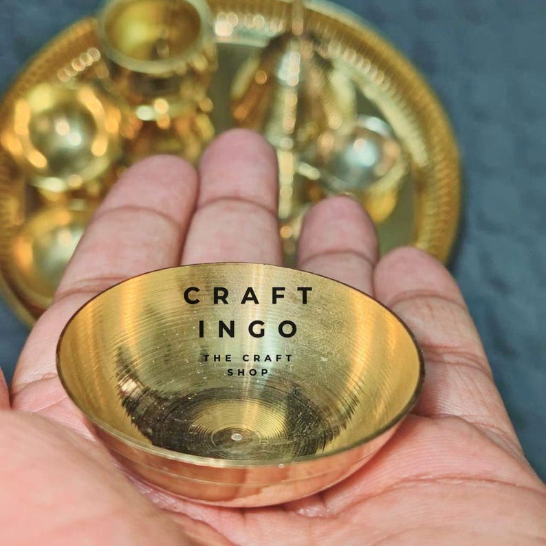 May include: A small, circular, polished brass bowl with a smooth, reflective interior surface. The text CRAFT INGO THE CRAFT SHOP is printed in black in the center of the bowl. A larger, matching brass tray with multiple circular indentations is visible.