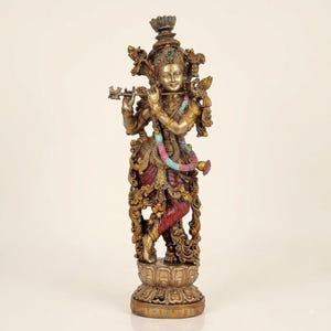 Puede incluir: Una estatua de tono bronce de una figura de pie tocando una flauta, adornada con un tocado ornamentado, joyas y una guirnalda de cuentas multicolor. La figura viste una prenda de color rojo oscuro y se apoya sobre una base de flor de loto.