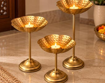 May include: Three gold-toned metal candle holders of varying heights, each featuring a flower-shaped bowl with a dimpled texture. Each holder stands on a circular base and contains a single lit tea light candle, casting a warm glow.