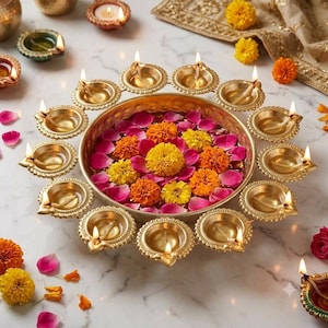 May include: A circular brass bowl filled with water, pink rose petals, and yellow and orange marigold flowers. The bowl is surrounded by a ring of small, lit brass oil lamps, known as diyas, arranged on a white marble surface.