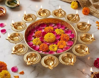 May include: A circular brass bowl filled with water, pink rose petals, and yellow and orange marigold flowers. The bowl is surrounded by a ring of small, lit brass oil lamps, known as diyas, arranged on a white marble surface.