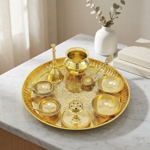 Könnte beinhalten: Ein poliertes Messing-Puja-Thali-Set mit einem großen runden Tablett mit komplizierten Blumengravuren. Das Set enthält eine hohe Glocke, einen kleinen Topf, zwei Öllampen, zwei kleine Schalen und einen Löffel auf der verzierten goldenen Oberfläche.