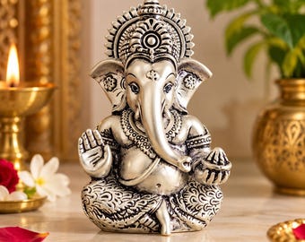 May include: A silver-toned Ganesha figurine featuring intricate embossed details on the crown, ears, and clothing. The deity is depicted in a seated position with one hand raised in a gesture of blessing and the other holding a small bowl.