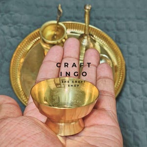 May include: A polished brass bowl held in a palm, with a matching brass tray, a small cup, and a bell visible in the background. The text CRAFT INGO THE CRAFT SHOP is overlaid on the image. The set features a metallic gold finish throughout.