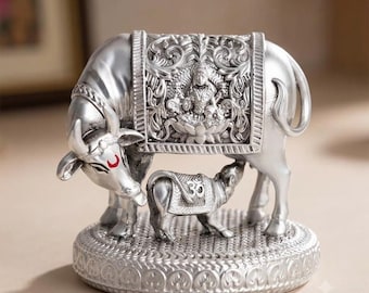 May include: A silver-toned metallic figurine of a cow and calf standing on a circular base. The cow features a red crescent mark on its forehead and a detailed blanket depicting a deity. The calf wears a blanket with the Om symbol embossed on it.