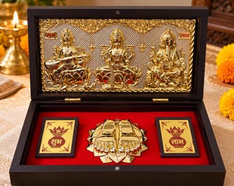 May include: A dark wooden box with a red velvet interior containing a gold-toned metal plaque featuring three deities, a central pair of gold feet on a lotus base, and two rectangular gold plates with the Hindi text Shubh and Labh.