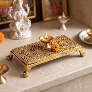 Puede incluir: Una plataforma rectangular de metal dorado con cuatro patas cortas, con patrones circulares de mandala en relieve y motivos de pétalos de loto. Una pequeña lámpara de aceite de latón con una llama encendida descansa sobre la superficie junto a una flor de caléndula naranja.
