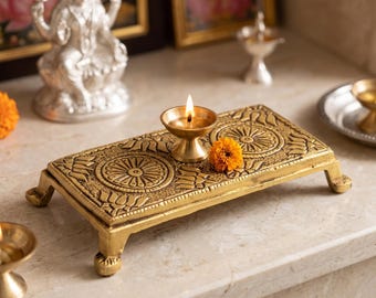 May include: A rectangular gold-toned metal platform with four short legs, featuring embossed circular mandala patterns and lotus petal motifs. A small brass oil lamp with a lit flame sits on the surface, accompanied by a single orange marigold flower.