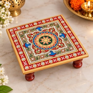 Könnte beinhalten: Ein quadratischer Holzständer mit einem handbemalten floralen Mandala-Design in Rot, Grün und Gold. Der Rand ist mit einem sich wiederholenden weißen Kreuzmuster und eingebetteten klaren Strasssteinen verziert. Er steht auf vier dunkelbraunen Füßen.