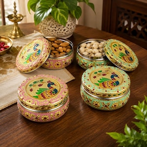 May include: Two round metal storage containers with lids, one pink and one light green, both featuring gold-tone embossed floral patterns and a central peacock motif. The containers are shown with their lids removed, revealing almonds and pistachios.