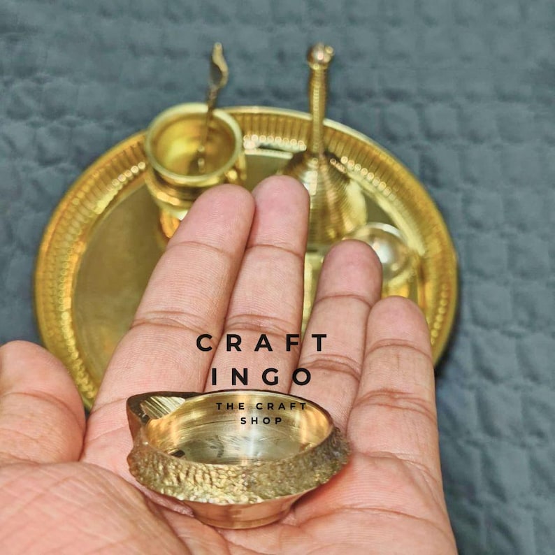 May include: A small, round, brass oil lamp with a textured outer rim held in the palm, with a larger brass tray in the background featuring a small brass cup and a bell-shaped object. The text CRAFT INGO THE CRAFT SHOP is visible in the center.