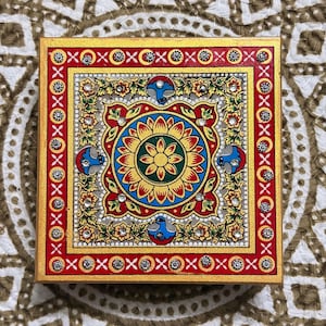 May include: A square decorative box featuring a central floral mandala design in yellow, red, and green. The border is painted red with white cross patterns and embedded clear rhinestones. Blue bird motifs are positioned around the centre.