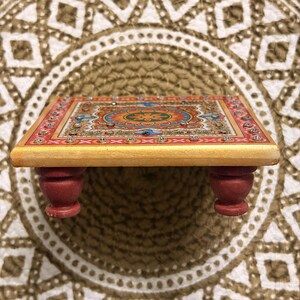 May include: A small, rectangular wooden platform with four short, rounded red legs. The top surface features a vibrant, multi-coloured floral mandala pattern in shades of orange, blue, and green, bordered by a red frame with embedded clear crystals.
