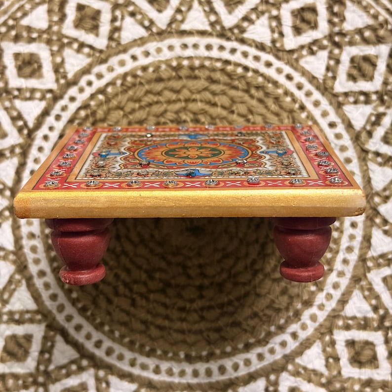 May include: A small, rectangular wooden platform with four short, rounded red legs. The top surface features a vibrant, multi-coloured floral mandala pattern in shades of orange, blue, and green, bordered by a red frame with embedded clear crystals.