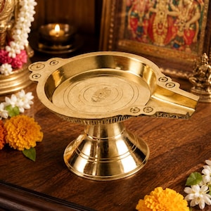 May include: A polished brass Abhishek Patra vessel featuring a wide circular basin with a prominent pouring spout, concentric etched rings on the interior base, and a sturdy pedestal stand, designed for ritual liquid offerings in a temple setting.