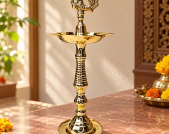 May include: A polished brass oil lamp featuring a tall, slender, ribbed pedestal base and a wide, circular reservoir. The top of the lamp is adorned with a decorative, stylized bird figure, creating a vertical, metallic structure.