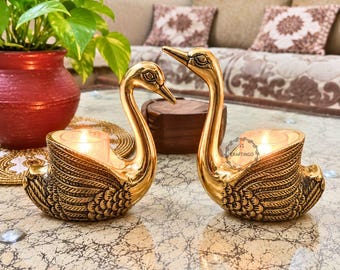 May include: Two gold-colored metal swan-shaped candle holders with textured wing details. Each swan features a circular opening on its back designed to hold a small tea light candle. The right swan has a small circular logo with the text CRAFTINGO.